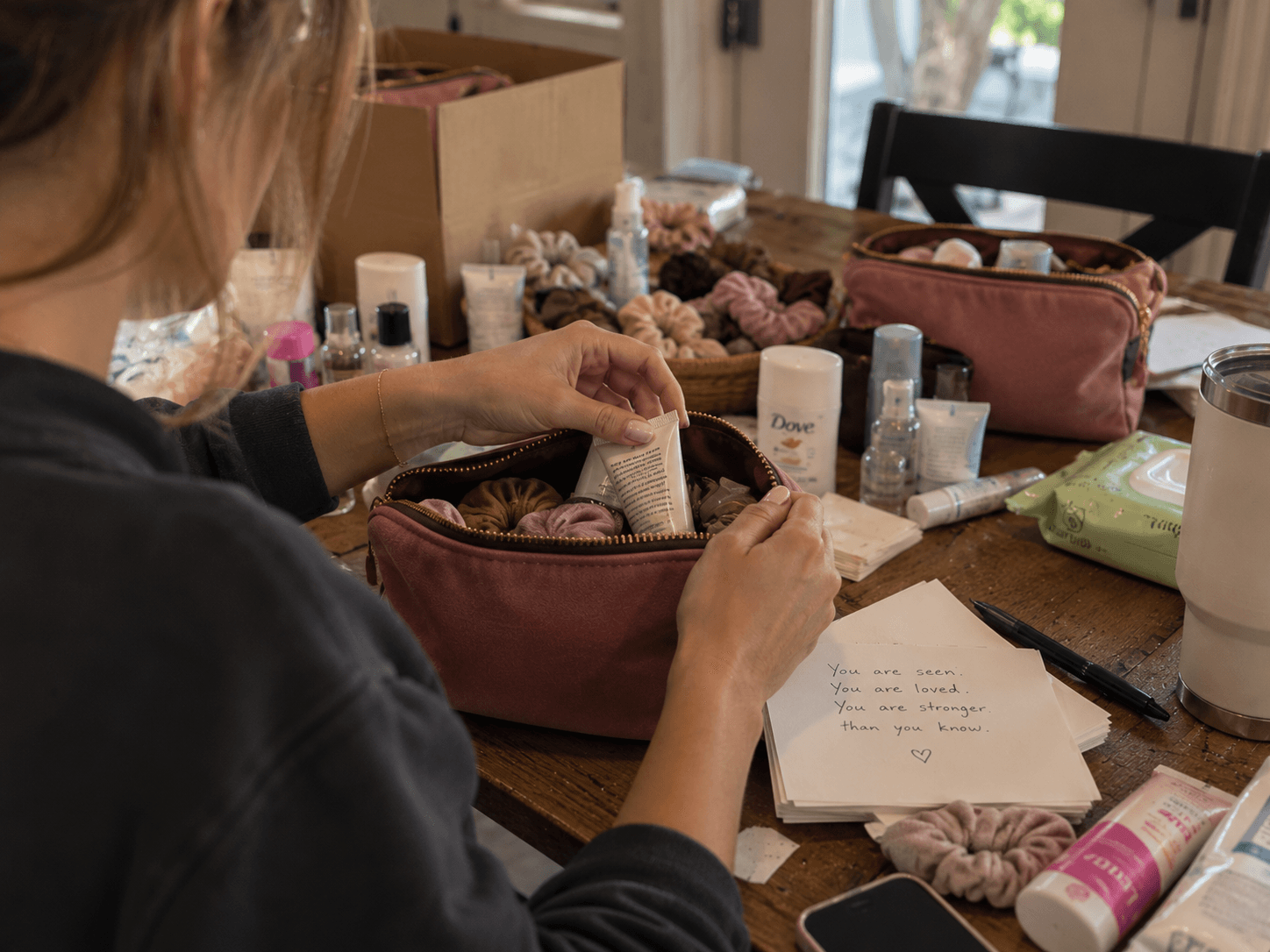 A woman tucking a hand cream into a pink accessory bag, with a handwritten note on the table beside her