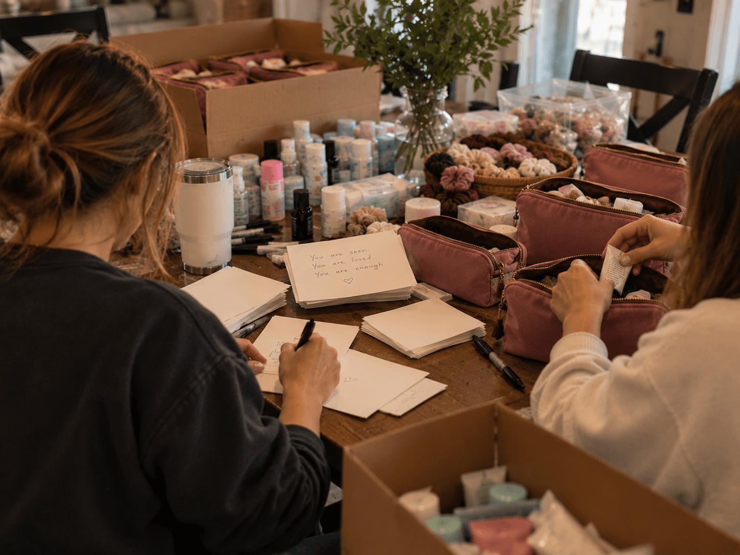 Two volunteers at a table, one writing notes and one filling pink accessory bags
